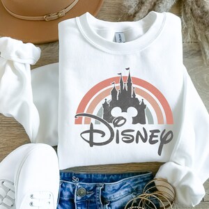 May include: White sweatshirt featuring a rainbow and a silhouette of Cinderella's castle with Mickey Mouse ears and the word "DISNEY" in black.
