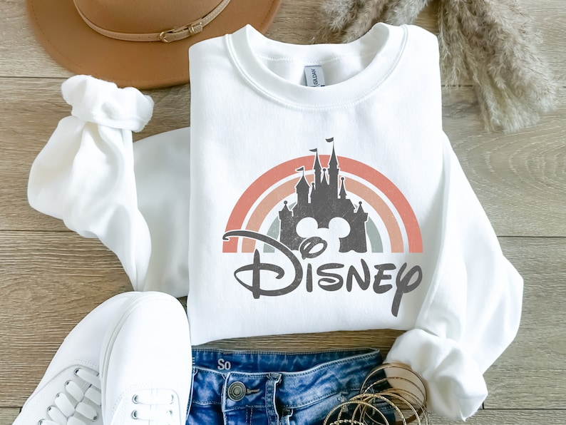 May include: White sweatshirt featuring a rainbow and a silhouette of Cinderella's castle with Mickey Mouse ears and the word "DISNEY" in black.