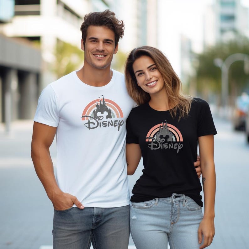May include: Two people wearing matching Disney t-shirts. One shirt is white, the other is black. Both shirts feature a rainbow design with a castle, Mickey Mouse silhouette, and the word "Disney".