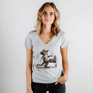May include: A light gray V-neck t-shirt featuring a cartoon cat wearing a cowboy hat and boots. The cat is walking with one leg raised, and a small cactus is visible in the background. The graphic is in shades of brown.