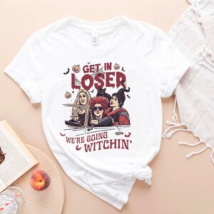 May include: White t-shirt with a graphic of three witches driving in a car. The text on the shirt reads "Get in Loser, We're Going Witchin'"