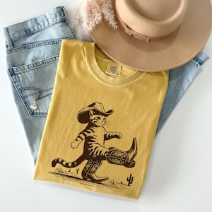 May include: Mustard yellow t-shirt featuring a cartoon cat wearing a cowboy hat and boots. The graphic is in brown and depicts the cat walking with a desert landscape and a cactus. A light brown hat and blue jeans are also in the image.