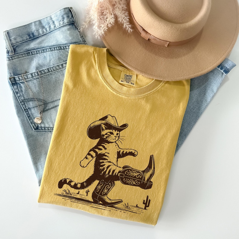May include: Mustard yellow t-shirt featuring a cartoon cat wearing a cowboy hat and boots. The graphic is in brown and depicts the cat walking with a desert landscape and a cactus. A light brown hat and blue jeans are also in the image.