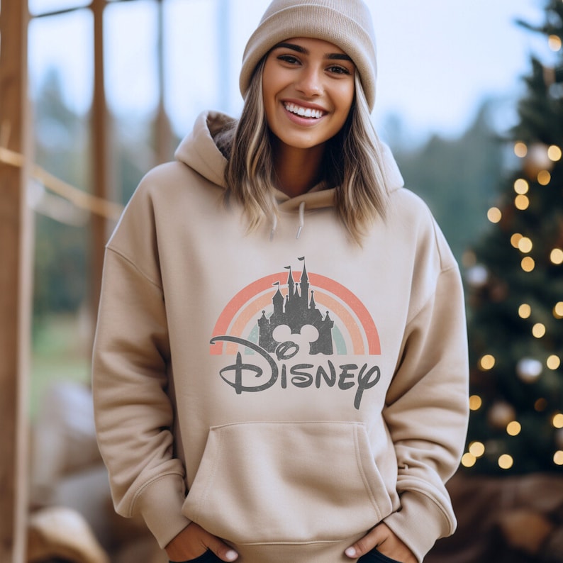 May include: A beige hoodie featuring a vintage-style rainbow and the text "DISNEY" with a silhouette of Cinderella's castle and Mickey Mouse ears.