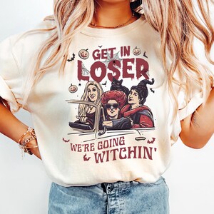 May include: A white t-shirt with a graphic of three women dressed as witches driving in a car. The text on the shirt reads "Get in Loser, We're Going Witchin'"
