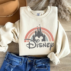May include: A cream-coloured sweatshirt featuring a graphic of a rainbow, Cinderella's castle, and Mickey Mouse ears with the word "DISNEY" underneath. The sweatshirt is folded and laid on a wooden surface with a brown hat and blue jeans.