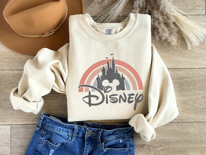 May include: A cream-coloured sweatshirt featuring a graphic of a rainbow, Cinderella's castle, and Mickey Mouse ears with the word "DISNEY" underneath. The sweatshirt is folded and laid on a wooden surface with a brown hat and blue jeans.