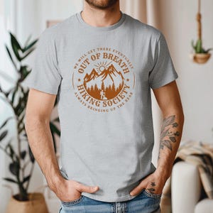 Może przedstawiać: Jasnoszara koszulka z okrągłym wzorem z napisem "OUT OF BREATH HIKING SOCIETY". Projekt zawiera pasmo g&oacute;rskie, słońce i drzewa w kolorze brązowym. Koszulka ma kr&oacute;tkie rękawy i okrągły dekolt.