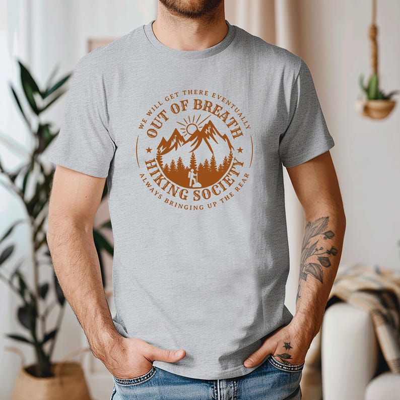 Może przedstawiać: Jasnoszara koszulka z okrągłym wzorem z napisem "OUT OF BREATH HIKING SOCIETY". Projekt zawiera pasmo g&oacute;rskie, słońce i drzewa w kolorze brązowym. Koszulka ma kr&oacute;tkie rękawy i okrągły dekolt.