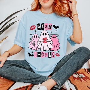 Pu&ograve; includere: Una maglietta blu chiaro con una grafica di tre fantasmi rosa. I fantasmi tengono un libro rosa e una chiave. Il testo "Mean Ghouls" &egrave; scritto in lettere nere con accenti rosa.