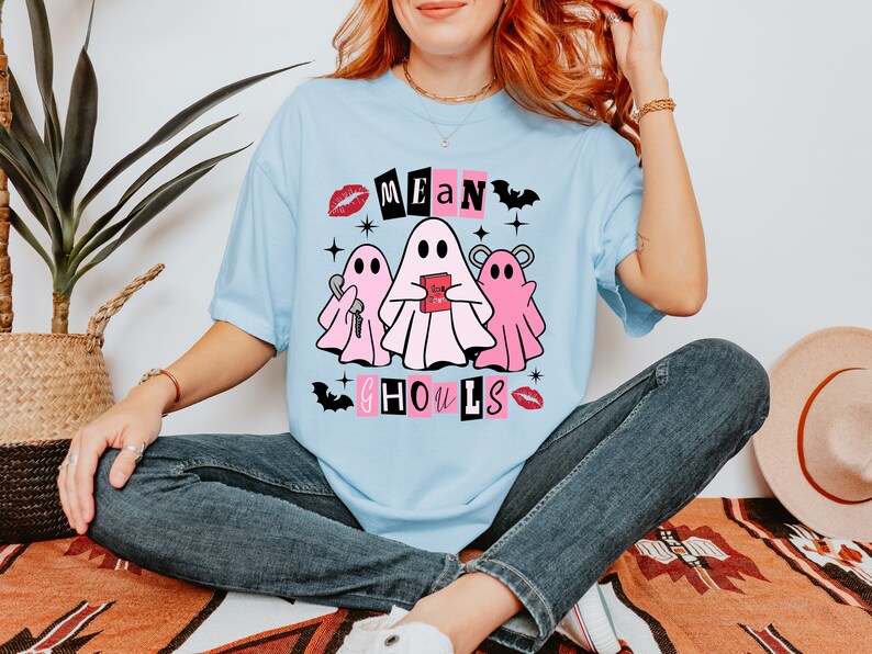 Pu&ograve; includere: Una maglietta blu chiaro con una grafica di tre fantasmi rosa. I fantasmi tengono un libro rosa e una chiave. Il testo "Mean Ghouls" &egrave; scritto in lettere nere con accenti rosa.