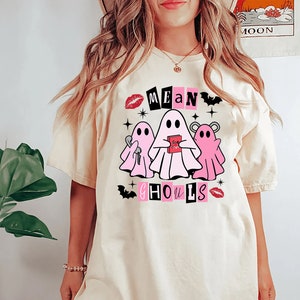 Pu&ograve; includere: Una maglietta bianca con un design grafico rosa e nero con tre fantasmi e il testo "Mean Ghouls".