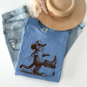 May include: A light blue t-shirt featuring a brown illustration of a cat wearing a cowboy hat and boots. The shirt is paired with light wash denim jeans and a tan hat. A gold necklace is visible in the jeans pocket.