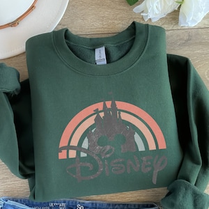 May include: A dark green sweatshirt featuring a vintage-style rainbow with the word "DISNEY" in a distressed font. The rainbow has a silhouette of Cinderella's castle in the centre.