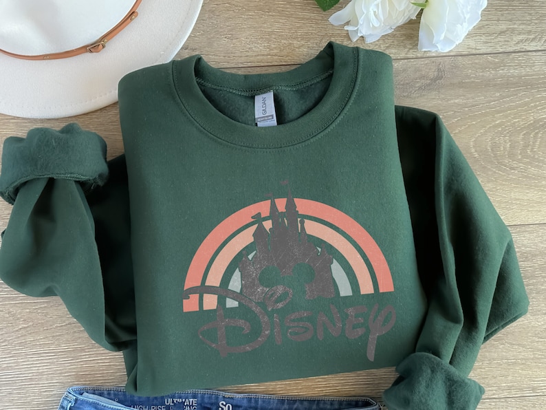 May include: A dark green sweatshirt featuring a vintage-style rainbow with the word "DISNEY" in a distressed font. The rainbow has a silhouette of Cinderella's castle in the centre.