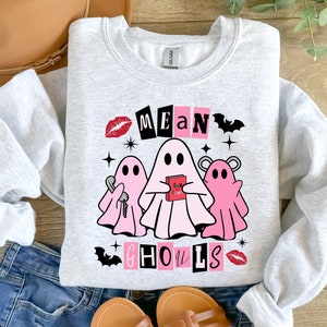 Pu&ograve; includere: Una felpa grigia con un design grafico rosa e nero con tre fantasmi e il testo "Mean Ghouls".