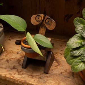 May include: A wooden planter shaped like a robot with two circular eye-like features and a central terracotta pot containing a green plant. The robot has a dark brown finish, angled legs, and a small arm-like protrusion on the side of the pot holder.