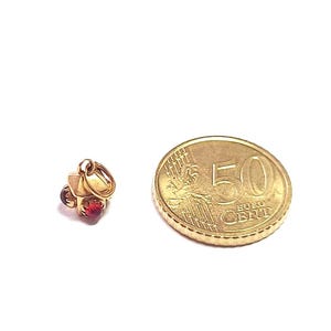 May include: A small gold-colored charm in the shape of a house with a red gemstone. Next to the charm is a 50 Euro cent coin. The charm has a loop for attaching to a necklace or bracelet.