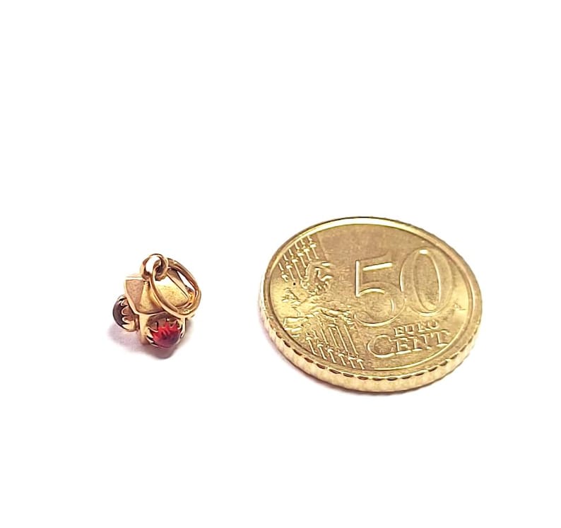 May include: A small gold-colored charm in the shape of a house with a red gemstone. Next to the charm is a 50 Euro cent coin. The charm has a loop for attaching to a necklace or bracelet.