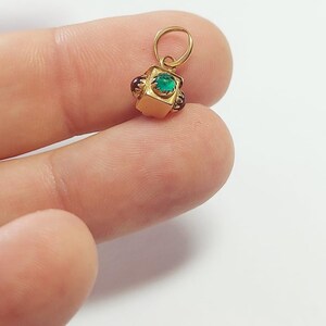 May include: A small, gold-colored pendant with a square, emerald-green gemstone at its center. The pendant has a loop at the top for a chain and is accented with small, dark red gemstones on each side. The pendant is held by a person's fingers.