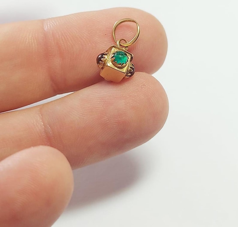 May include: A small, gold-colored pendant with a square, emerald-green gemstone at its center. The pendant has a loop at the top for a chain and is accented with small, dark red gemstones on each side. The pendant is held by a person's fingers.