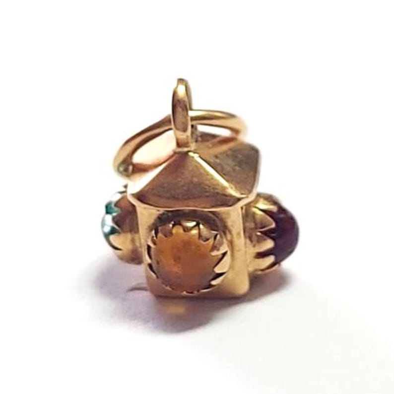 May include: A gold-colored charm in the shape of a lantern. The charm has a small loop at the top for attachment. It features a brown stone and two smaller stones in teal and burgundy. The lantern has a decorative sunburst design.