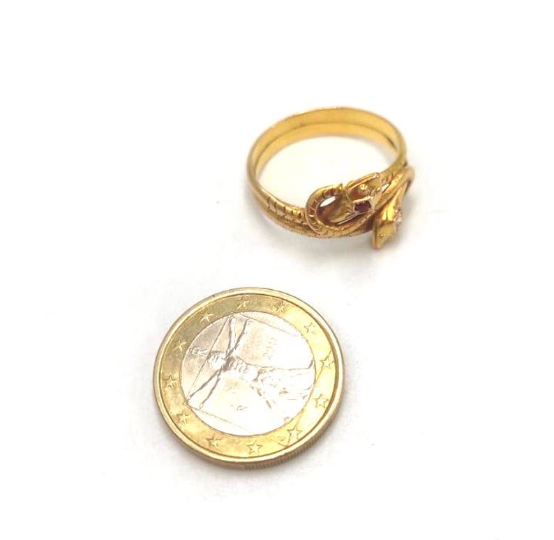 May include: A gold-colored ring in the shape of a snake, with a small red stone and a clear stone. A Euro coin is next to the ring for size comparison. The ring is on a white background.