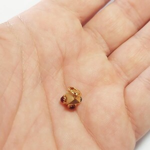 May include: A small, gold-colored charm with a geometric design. The charm has a central, multi-faceted shape with small, dark red and blue accents. It features a small loop for attaching to a necklace or bracelet.