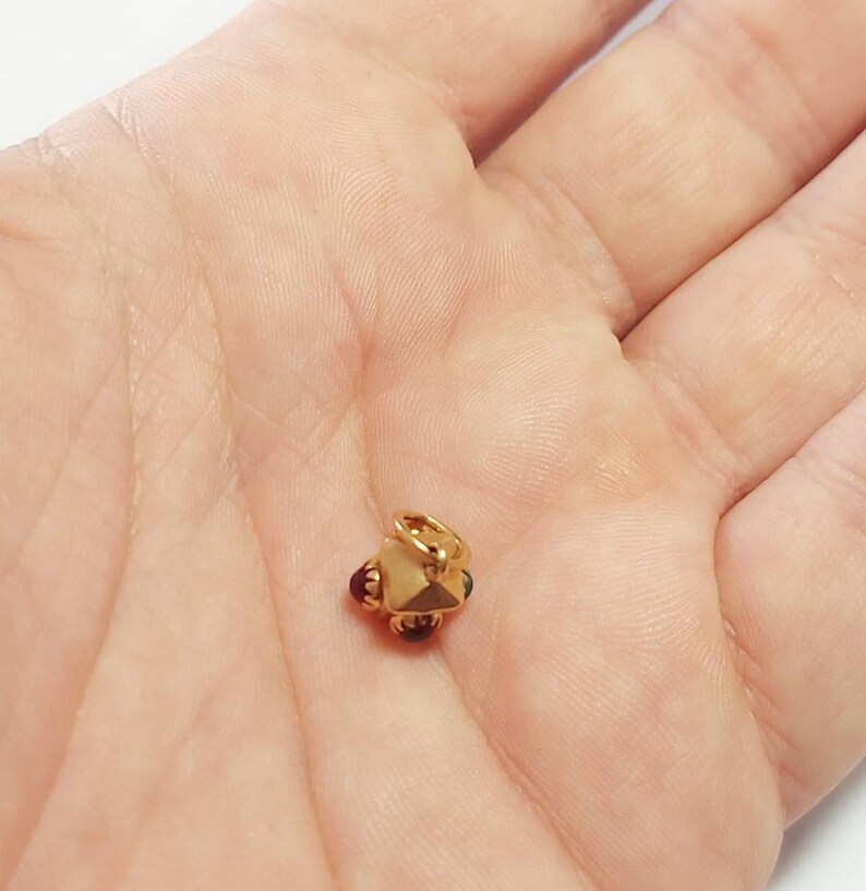 May include: A small, gold-colored charm with a geometric design. The charm has a central, multi-faceted shape with small, dark red and blue accents. It features a small loop for attaching to a necklace or bracelet.