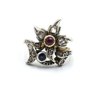 May include: A vintage silver ring with a floral design. The ring features a central ruby gemstone surrounded by smaller diamonds and a blue sapphire. The ring has a gold band and is set against a white background.
