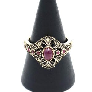 Könnte beinhalten: Verzierter goldfarbener Ring mit einem zentralen ovalen Rubin-Edelstein und kleineren Rubin-Akzenten. Der Ring hat ein filigranes Design mit aufwendigen Details. Der Ring wird auf einem schwarzen kegelförmigen Ständer vor weißem Hintergrund präsentiert.