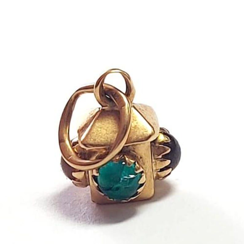 May include: A gold-colored charm with a house-like design. The charm features a vibrant green gemstone and two smaller, dark-colored stones. The charm has a loop for attaching to a necklace or bracelet. The charm is likely made of gold.