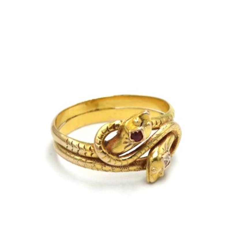 May include: A gold-toned ring in the shape of two intertwined snakes. The snakes have textured scales and ruby and diamond accents. The ring is set against a white background.
