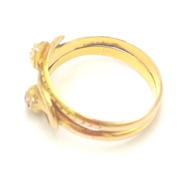 May include: A gold-toned ring with two small, round gemstones. The ring has a unique, intertwined design, with a smooth, polished finish. The gemstones are set in decorative settings, adding a touch of elegance. The ring is likely made of gold or a gold-colored metal.