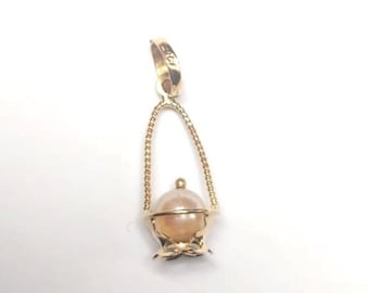 Pode incluir: Pingente dourado com uma pérola de cor pêssego claro em um design de cesta decorativa. A cesta tem uma alça em forma de laço e uma base floral. O pingente é projetado para joias.