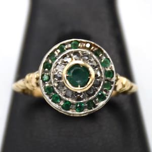 Puede incluir: Un anillo de oro con una piedra preciosa verde redonda en el centro, rodeada por un halo de pequeñas piedras claras y un borde exterior de pequeñas piedras verdes. El engaste es circular con una banda de oro amarillo de textura trenzada.