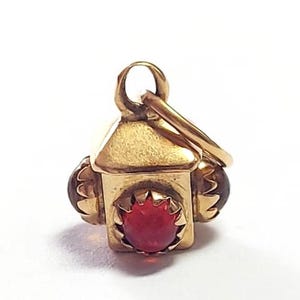 May include: A gold-colored lantern charm with a red gemstone at the center. The charm has a small loop at the top for attaching to a chain or bracelet. Two brown gemstones are on the sides of the lantern.