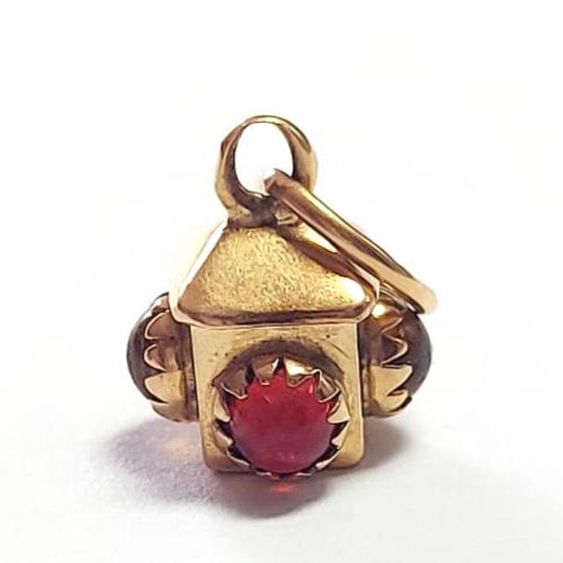 May include: A gold-colored lantern charm with a red gemstone at the center. The charm has a small loop at the top for attaching to a chain or bracelet. Two brown gemstones are on the sides of the lantern.