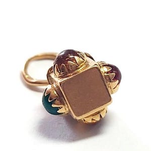 May include: A gold-colored nose ring with a square center and three surrounding gemstones. The gemstones are a mix of red and teal. The nose ring is set against a white background.