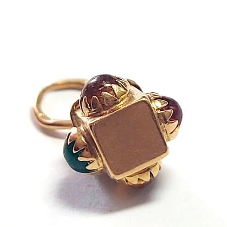 May include: A gold-colored nose ring with a square center and three surrounding gemstones. The gemstones are a mix of red and teal. The nose ring is set against a white background.