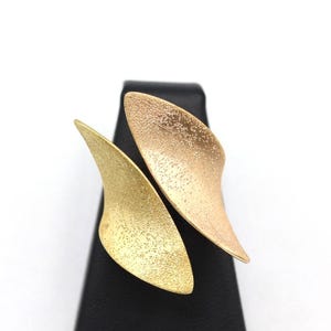 May include: A gold-coloured ring with a textured surface. The ring features two curved, leaf-like shapes that overlap. The ring is displayed on a black stand against a white background.