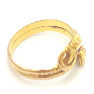 May include: A gold-colored ring with a decorative knot design and small red gemstones. The ring has a double band and a textured pattern along the lower portion. The ring is set against a white background.