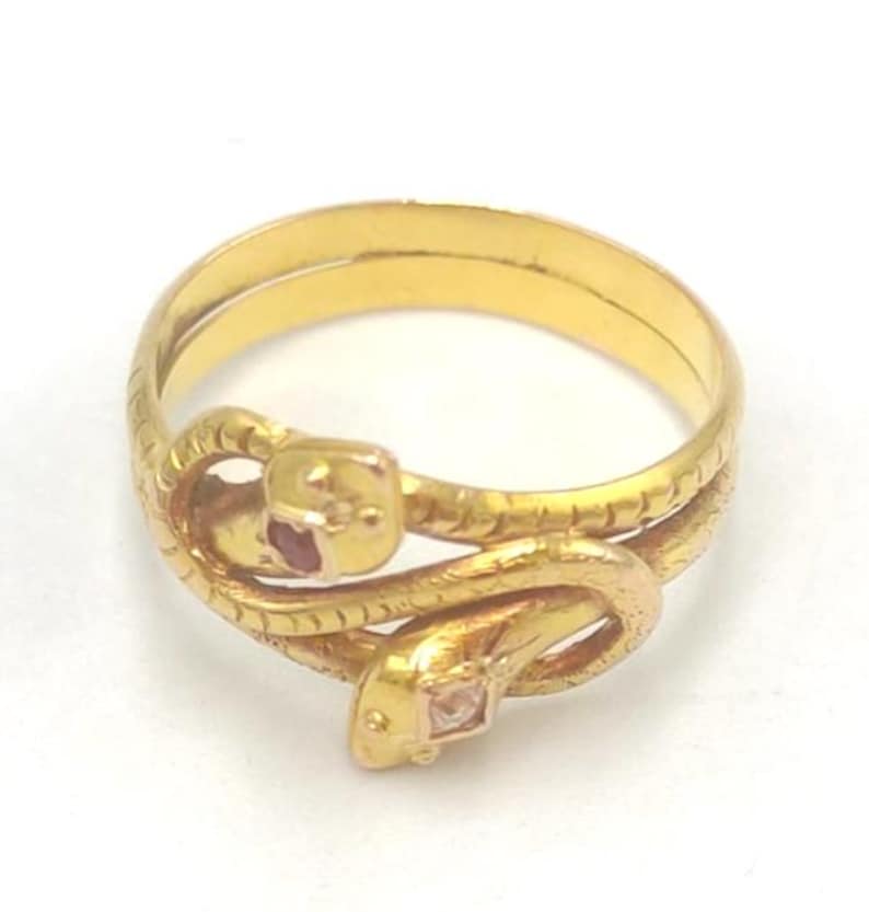 May include: A gold-colored ring featuring a snake design. The ring has two snakes intertwined, with one snake's head adorned with a red gemstone and the other with a clear gemstone. The ring is set against a white background.