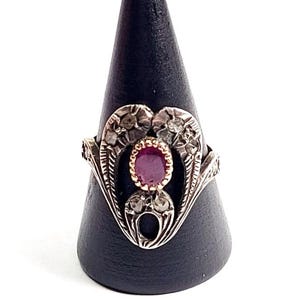 Op de afbeelding: Een antieke zilveren ring met een ovale robijn edelsteen in het midden. De ring heeft ingewikkelde bloemen- en bladerenontwerpen, met kleinere accentstenen rondom de robijn. De ring wordt getoond op een zwarte kegelvormige standaard.