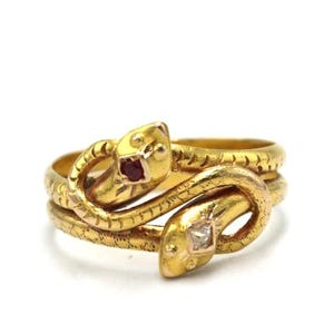 May include: A gold-toned ring featuring a double snake design. Each snake head has a gemstone eye, one red and one clear. The ring coils around the finger, creating a unique and eye-catching piece of jewelry.