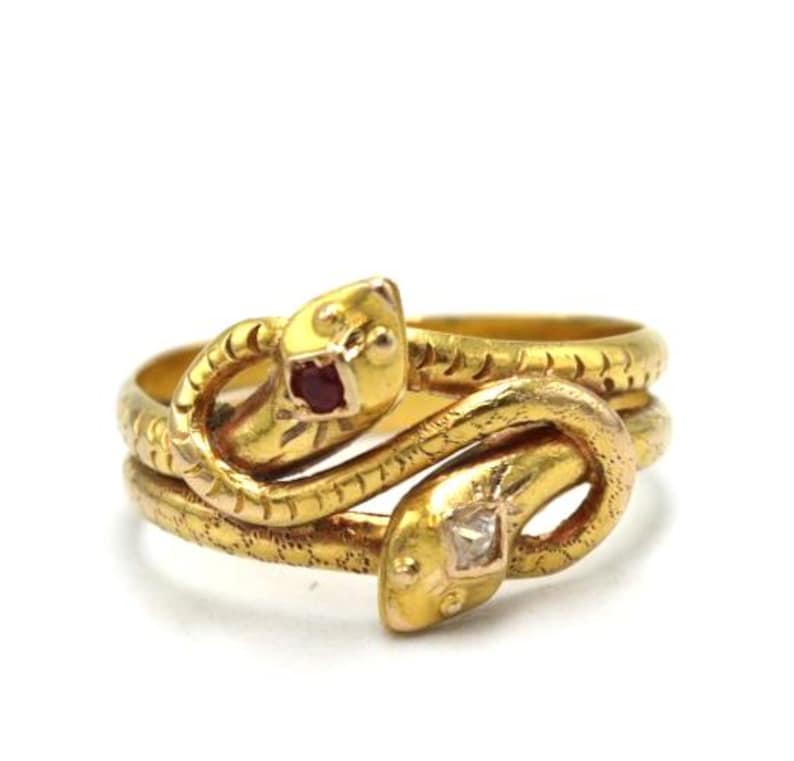 May include: A gold-toned ring featuring a double snake design. Each snake head has a gemstone eye, one red and one clear. The ring coils around the finger, creating a unique and eye-catching piece of jewelry.