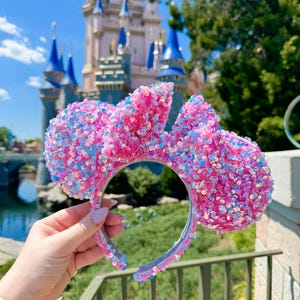 Pode incluir: Tiara de orelhas de rato rosa com lantejoulas e detalhes iridescentes. As orelhas são grandes e fofas, com uma tiara transparente. O fundo apresenta um castelo e vegetação.