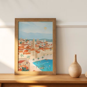 Puede incluir: Una ilustración enmarcada de una ciudad costera con edificios naranjas, palmeras y una masa de agua azul. El marco es de madera y tiene un acabado marrón claro.
