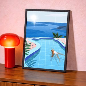 May include: A framed art print shows a person swimming in a blue pool, with a blue ocean and sky in the background. The artwork has a black frame and is displayed on a wooden surface next to a red lamp.
