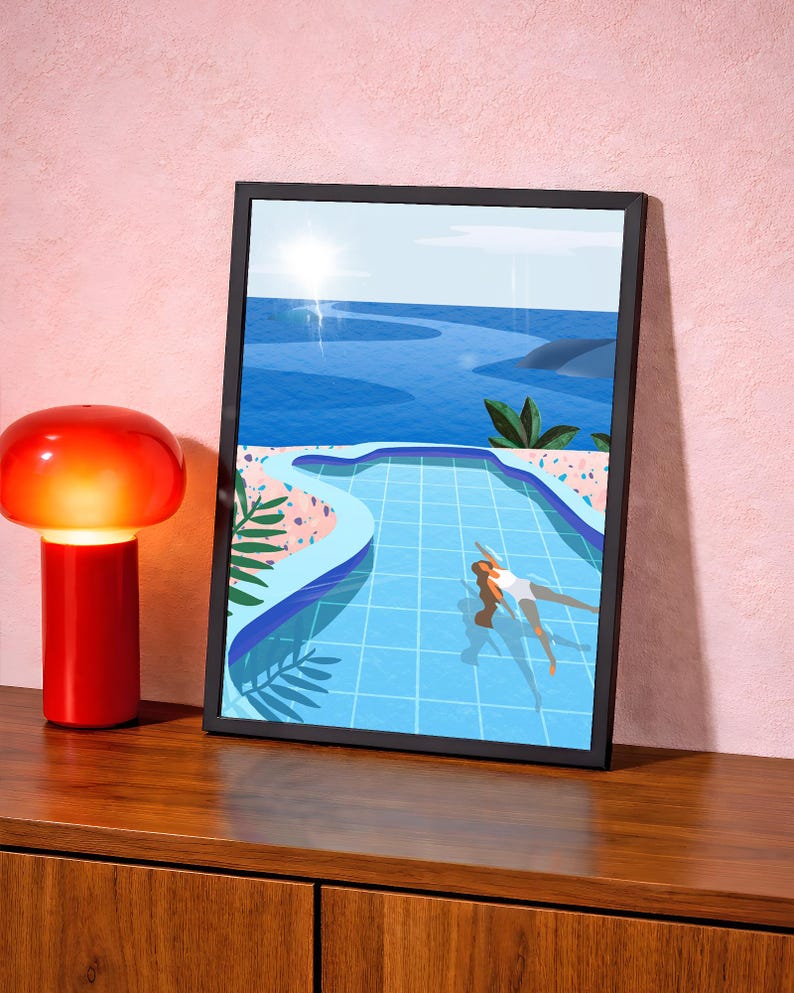 May include: A framed art print shows a person swimming in a blue pool, with a blue ocean and sky in the background. The artwork has a black frame and is displayed on a wooden surface next to a red lamp.